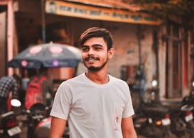 A young Indian man smiling outdoors in casual white attire on a bustling street.